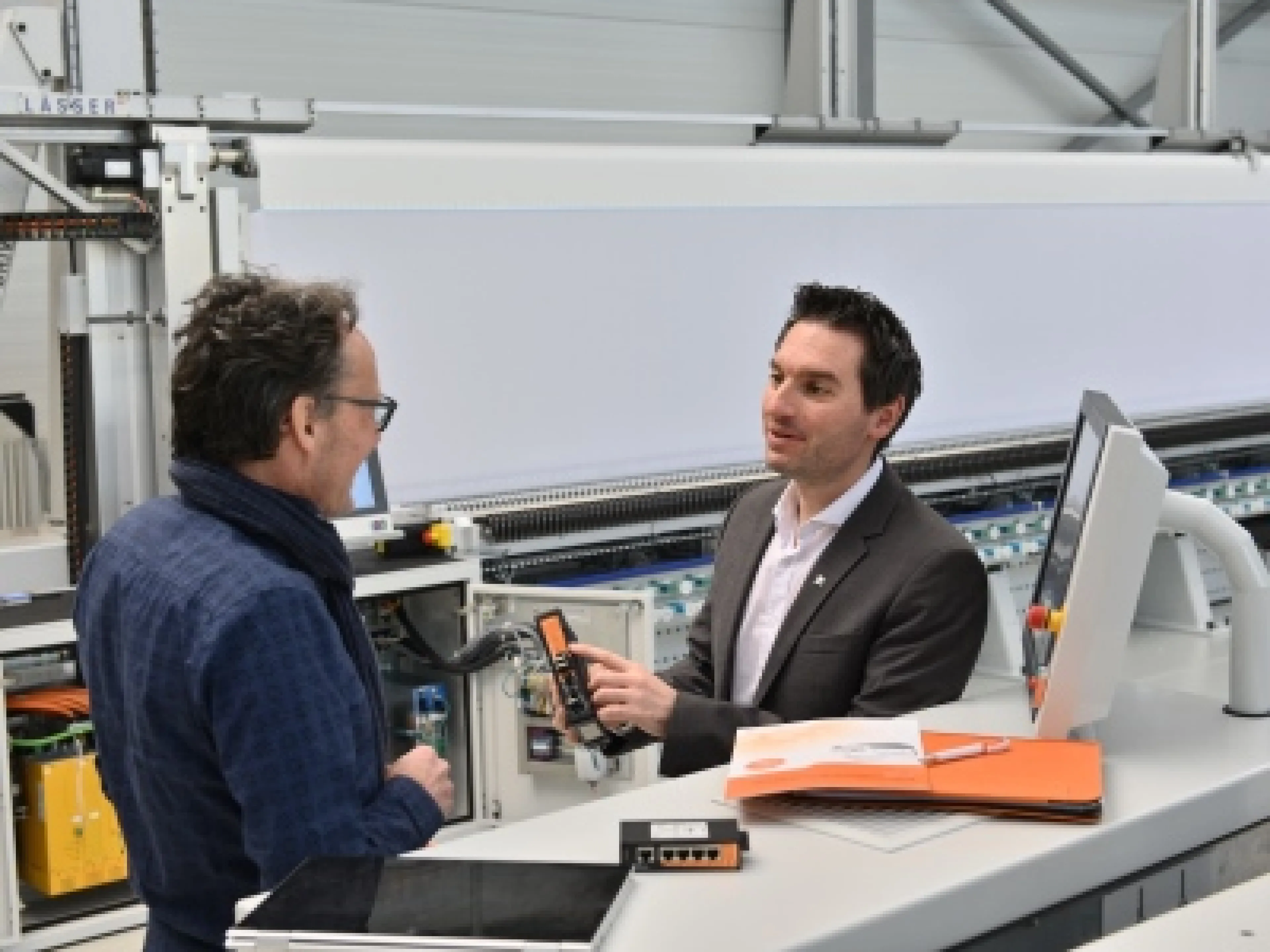 Vor der Stickmaschine MVD71 erklärt Peter Hirt, Segment Manager Machinery & Automation Sales, Patrik Schwarz, Softwareentwickler und Projektverantwortlicher bei Lässer, die Vorteile des Industrial Security Routers von Weidmüller